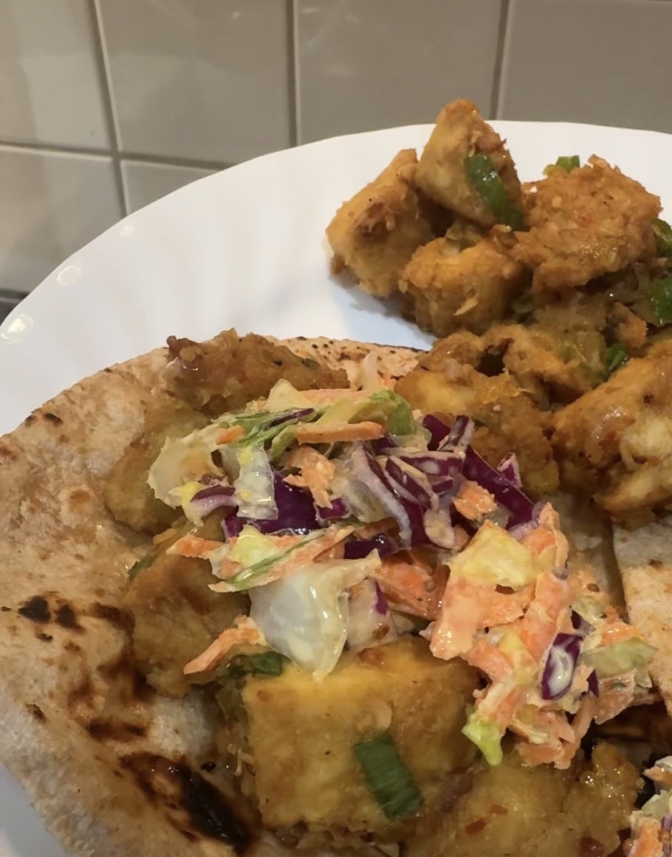 Crispy orange tofu in a charred flour tortilla, topped with shredded purple cabbage, green cabbage and carrot slaw, with a plate of glazed tofu pieces and spring onion behind it.