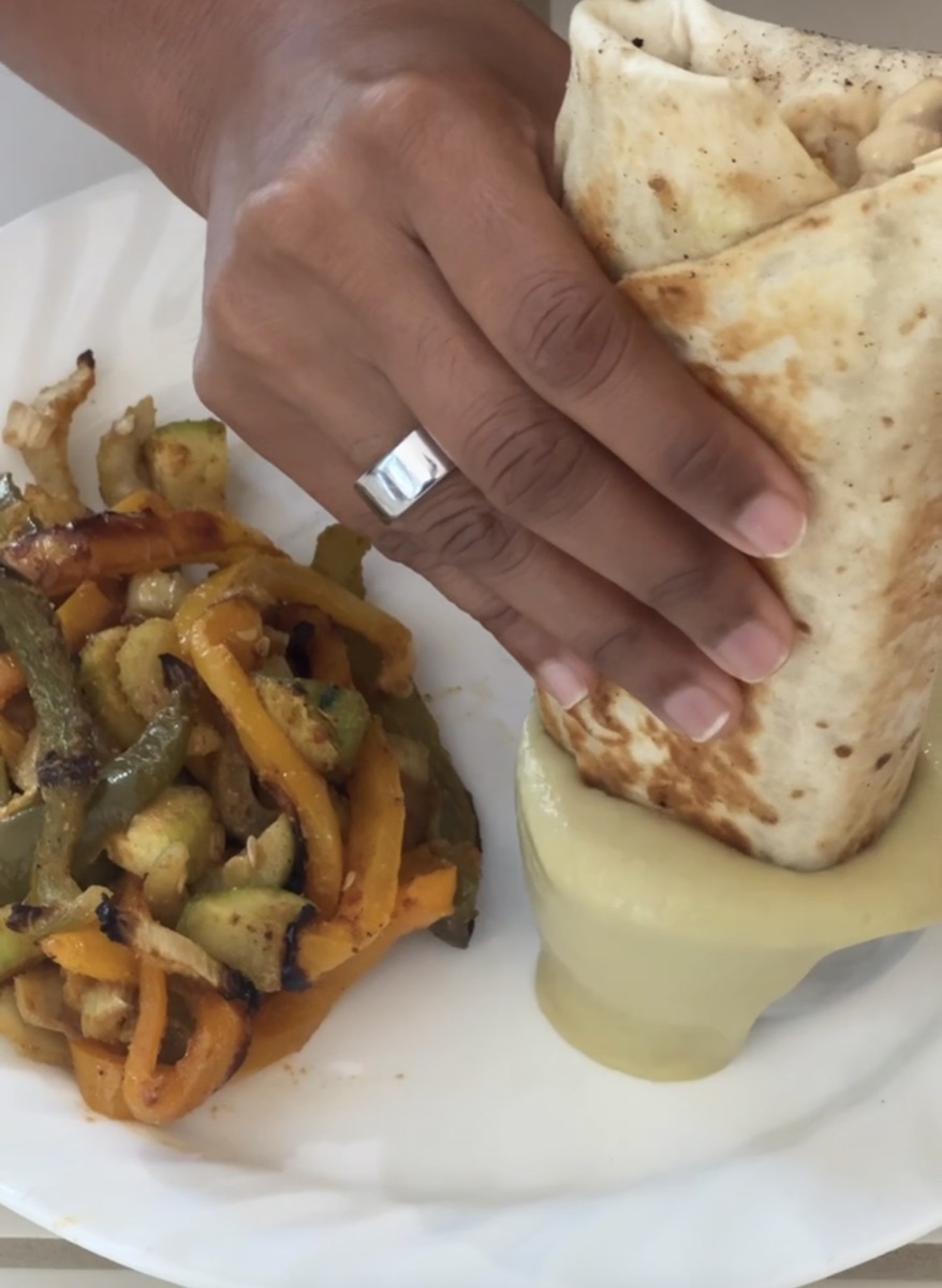 A seared high-protein bean and cheese burrito being dipped into salsa verde, with charred air-fried fajita veggies - peppers, zucchini and onion - on the plate alongside.